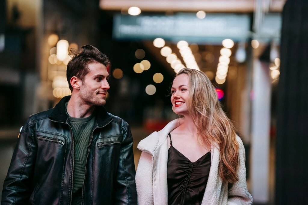 Positive couple in stylish clothes walking on street against building with glowing lights looking at each other in daytime