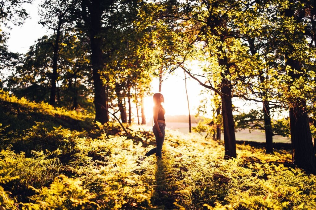 Woman walking in a sunlit forest during golden hour, capturing nature's beauty.