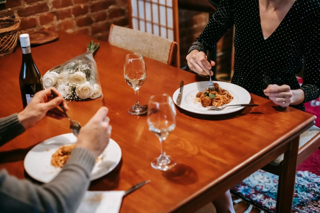 Crop faceless couple sitting in chairs at table while having dinner with pasta with meatballs on plates near cutlery and bott