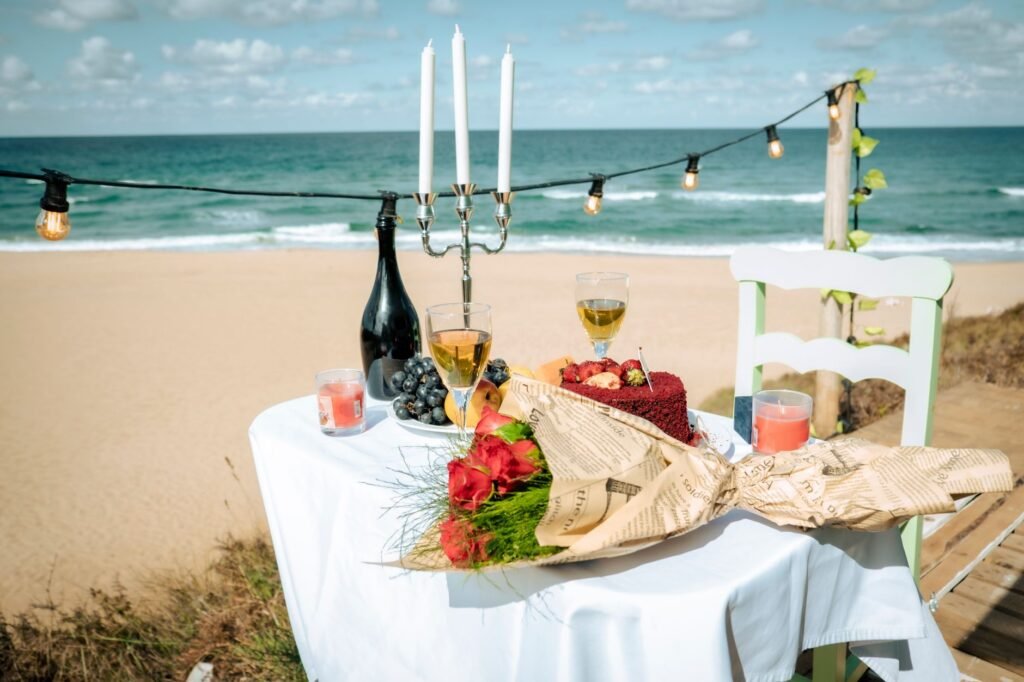 Romantic beachfront setup with champagne, cake, and roses under string lights.