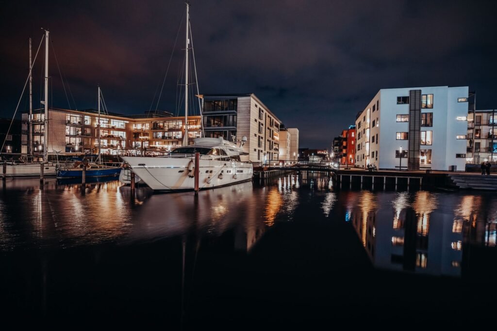 A serene night view of Odense marina featuring illuminated buildings and moored boats reflecting in calm waters.
