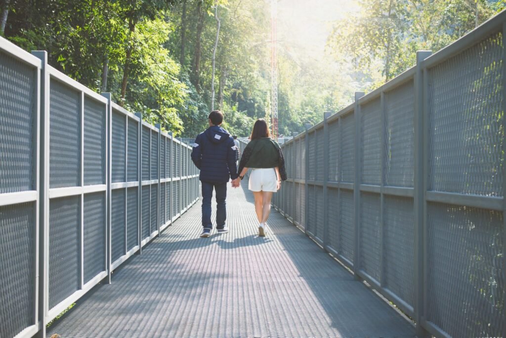 A couple holding hands walking along a forest bridge surrounded by lush greenery.