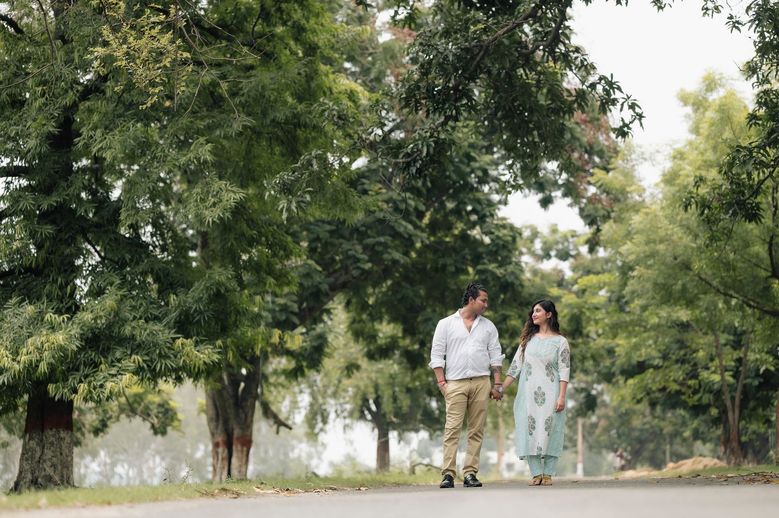 A romantic couple walks hand in hand through a vibrant green park, surrounded by trees.