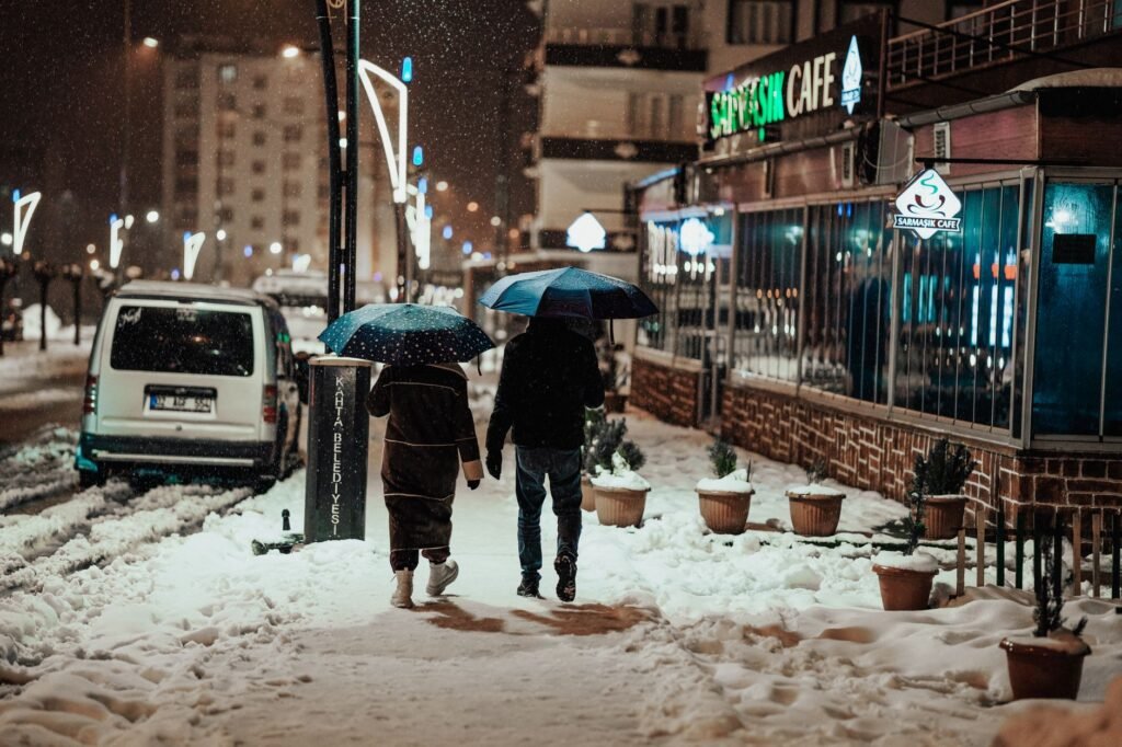 A couple with umbrellas walking on a snowy city street at night near a cozy cafe.