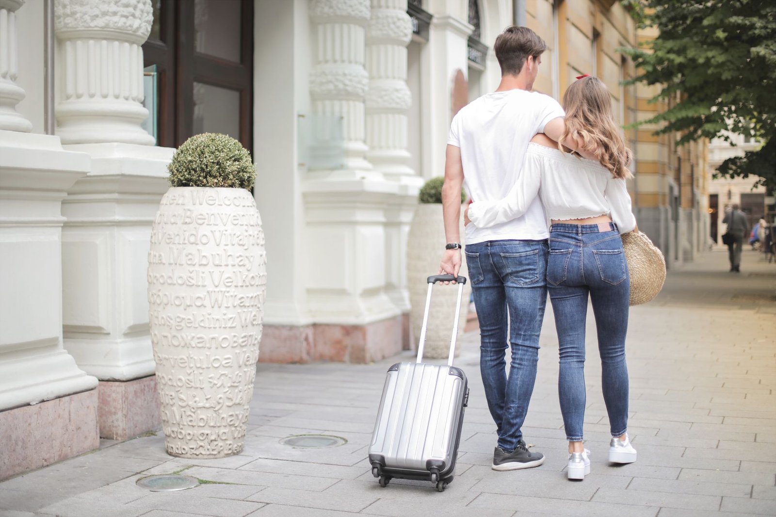 Back view of traveling couple in love wearing casual clothes walking with luggage and hugging while strolling along sidewalk