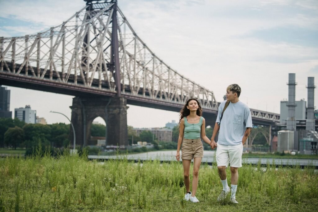 Asian man holding dreamy ethnic beloved by hand while strolling on grass near city bridge