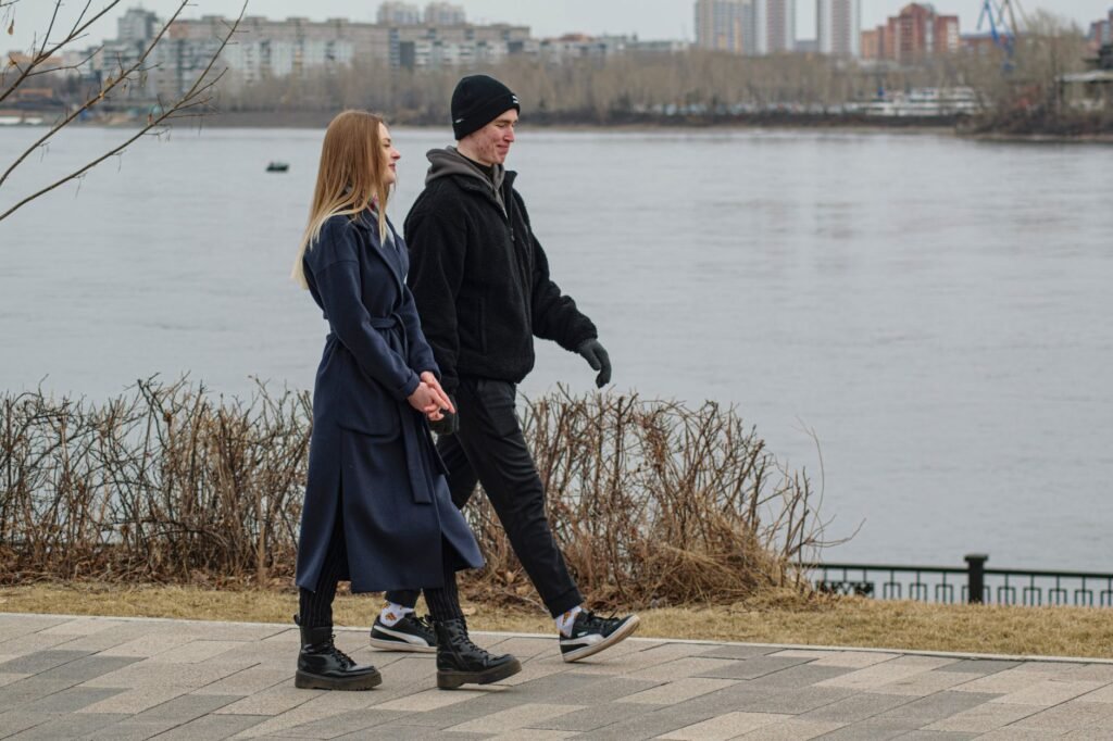 A young couple enjoys a winter walk along a riverside path in an urban park setting.