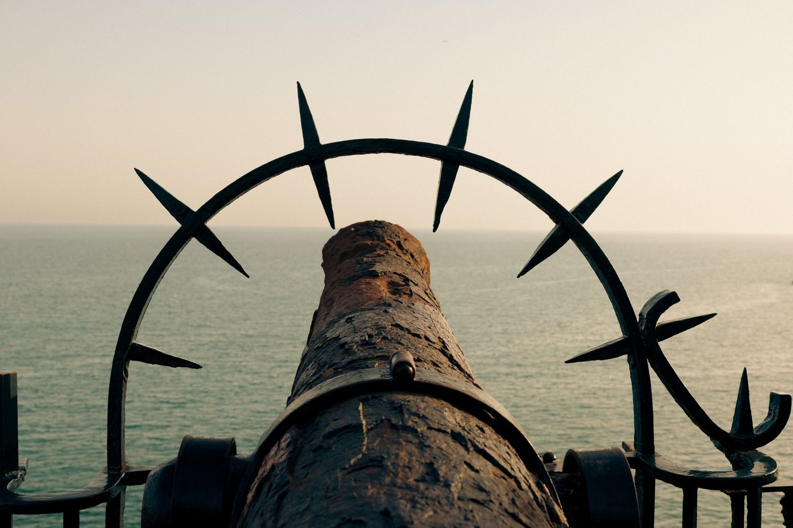 A rusted cannon aimed towards the sea, framed by metal spikes, suggesting a historic maritime setting.