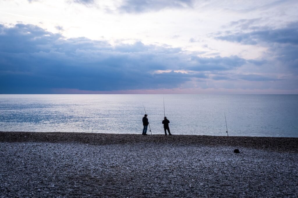 Two people fishing on a rocky beach at twilight, with calm sea and cloudy sky.