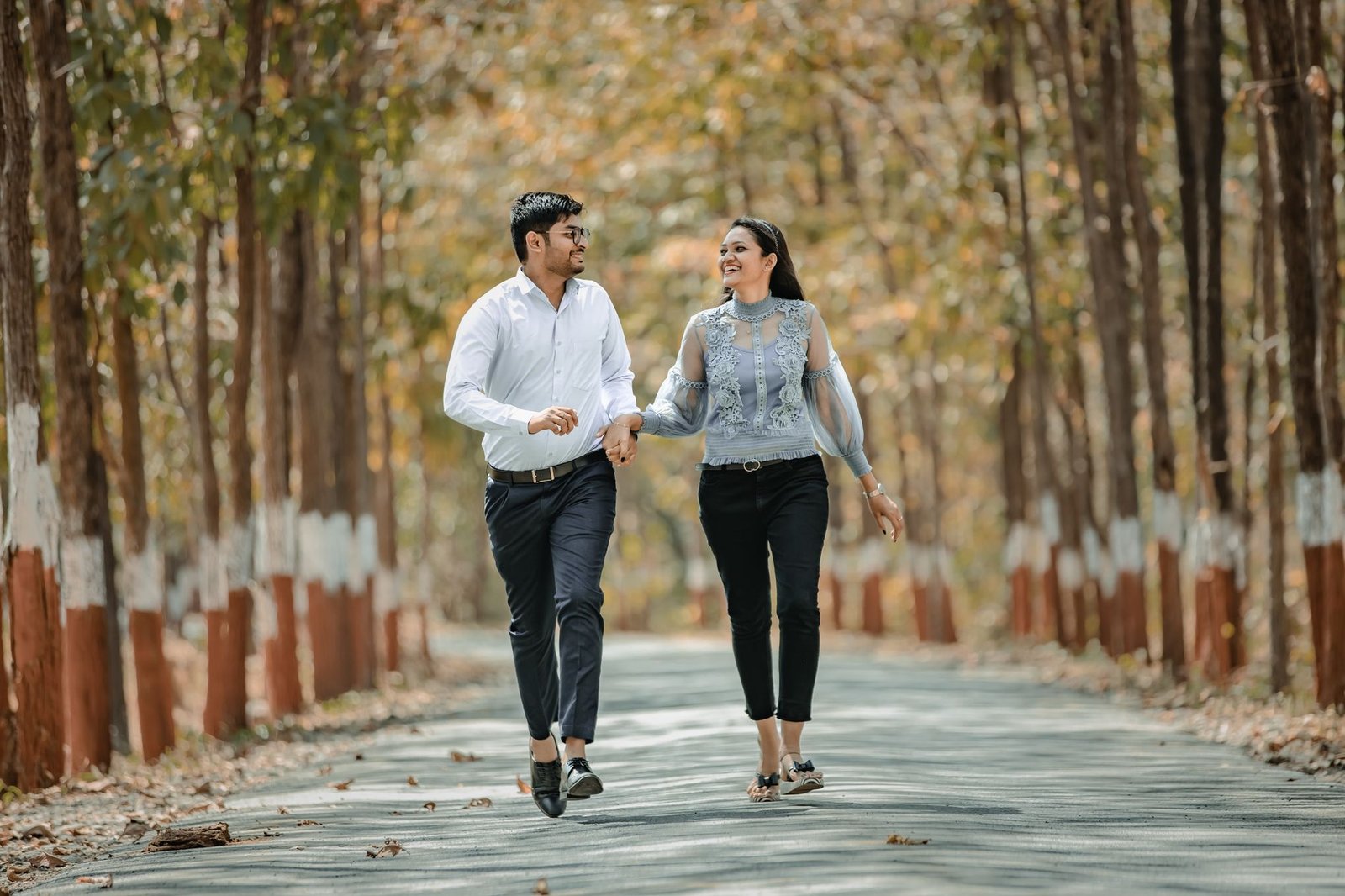 Smiling couple holding hands and walking down a tree-lined road, enjoying a sunny day in the park.