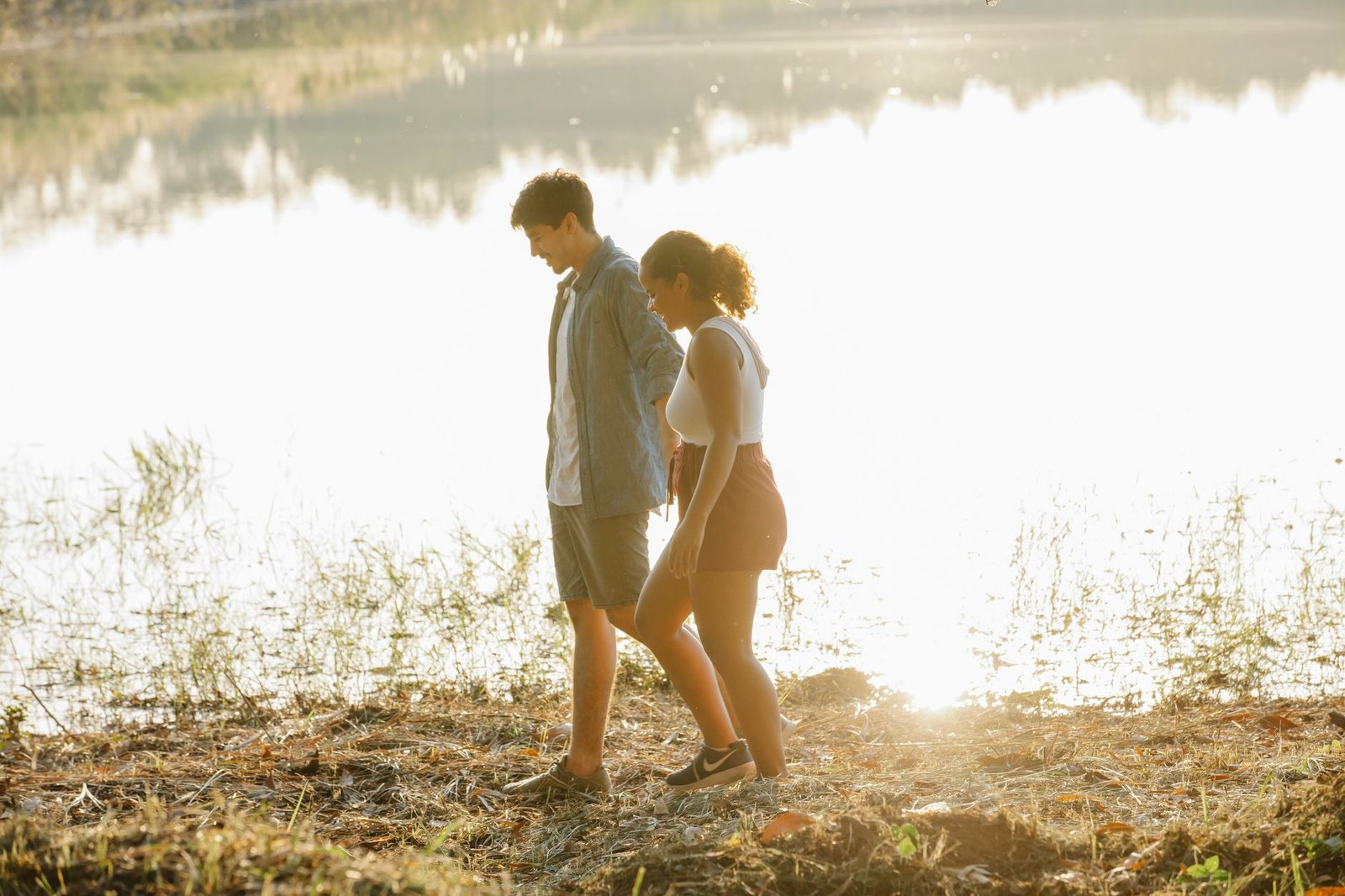 Side view of positive young multiracial couple in casual clothes holding hands while walking on grassy coast of calm lake dur