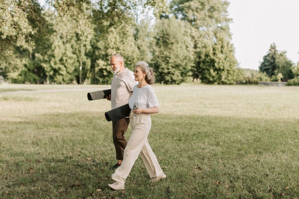 Elderly couple walking on grass carrying yoga mats, enjoying a sunny day outdoors.
