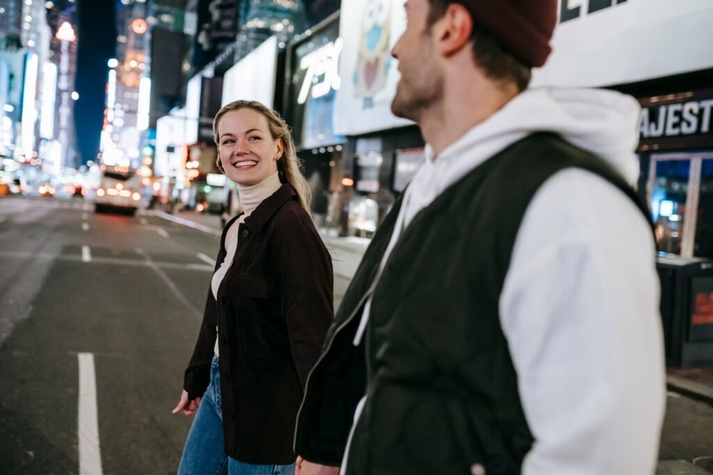 Cheerful man and woman smiling and crossing asphalt roadway with cars while walking on street with glowing buildings in eveni