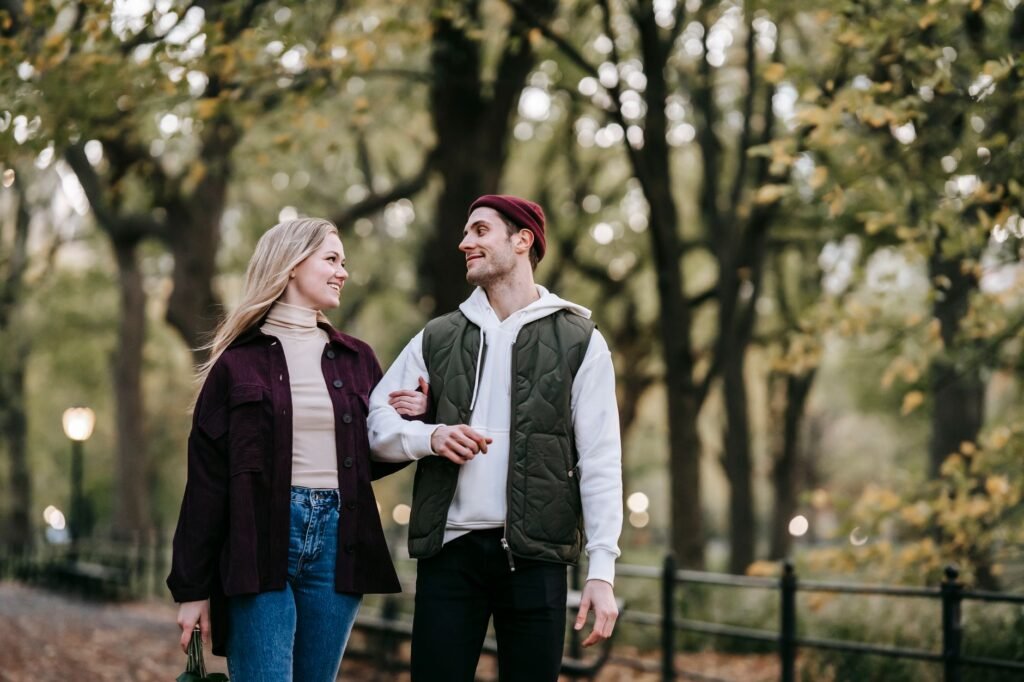 Cheerful couple smiling and walking in park with green trees and lights on blurred background