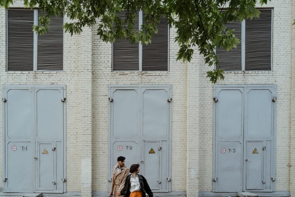 Couple walking hand in hand by an industrial building with large metal doors and a tree overhead.
