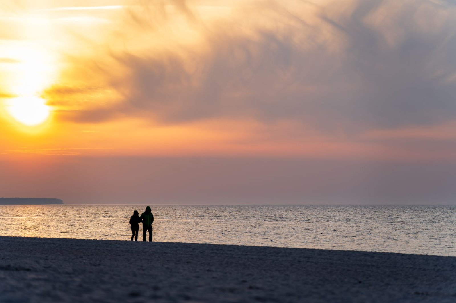 Couple walking along Kołobrzeg beach at sunset, embraced by a vivid, colorful sky.
