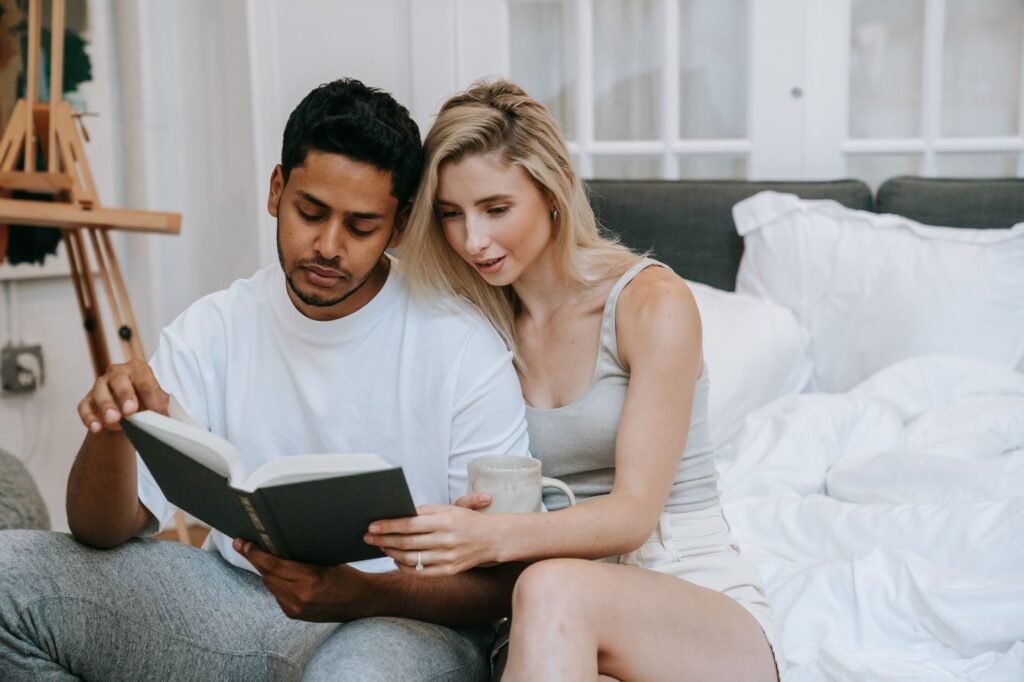 A young couple in pajamas reading a book together on a cozy bedroom bed, enjoying a relaxed indoor morning.