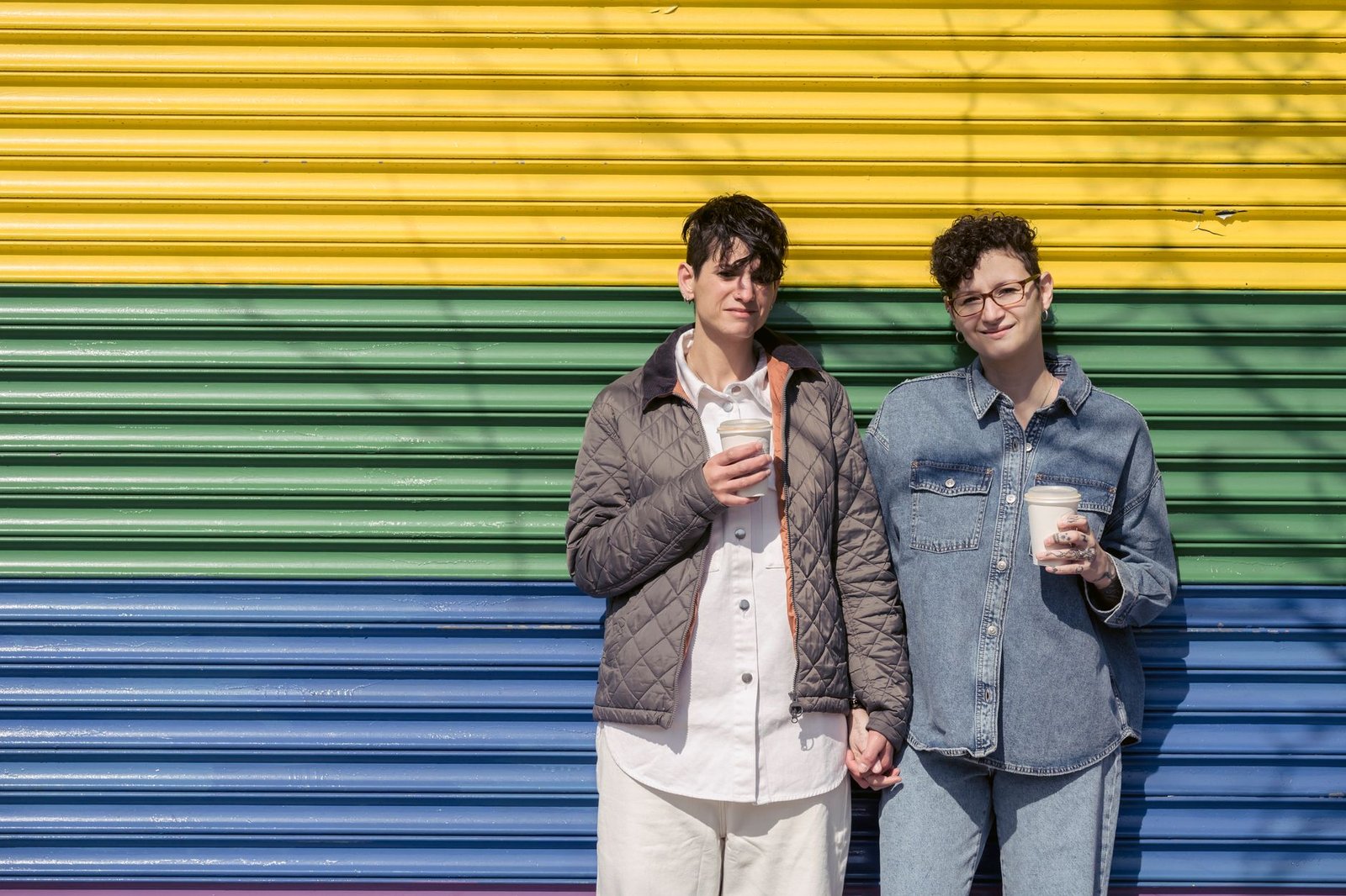 Trendy young lesbian couple with similar stylish haircut in casual clothes holding hands and drinking takeaway coffee near pr