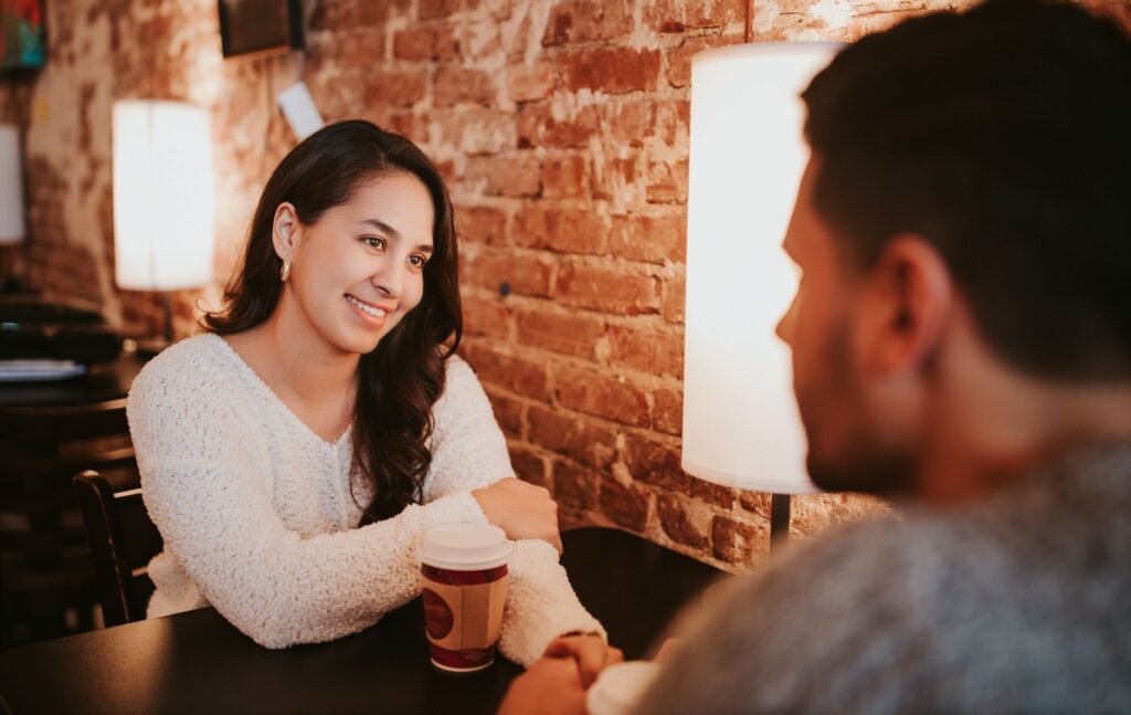 Young couple in casual outfit sitting at table in cafe with cups of drink on date near brick wall and holding hands