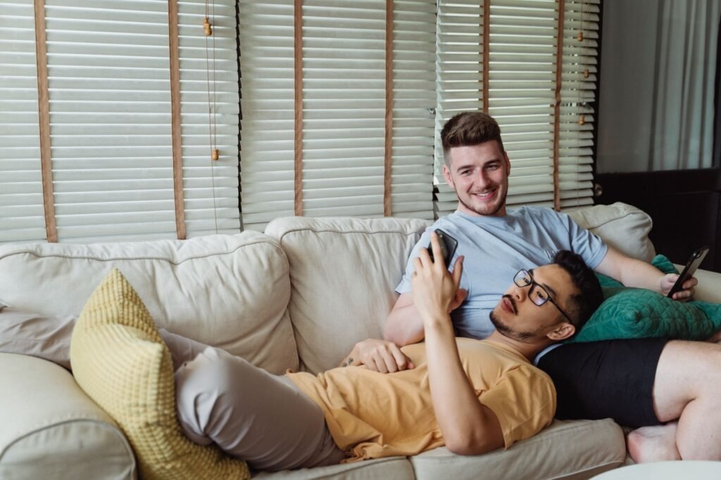 Casual scene of an interracial gay couple lounging on a sofa, enjoying their smartphones with smiles.