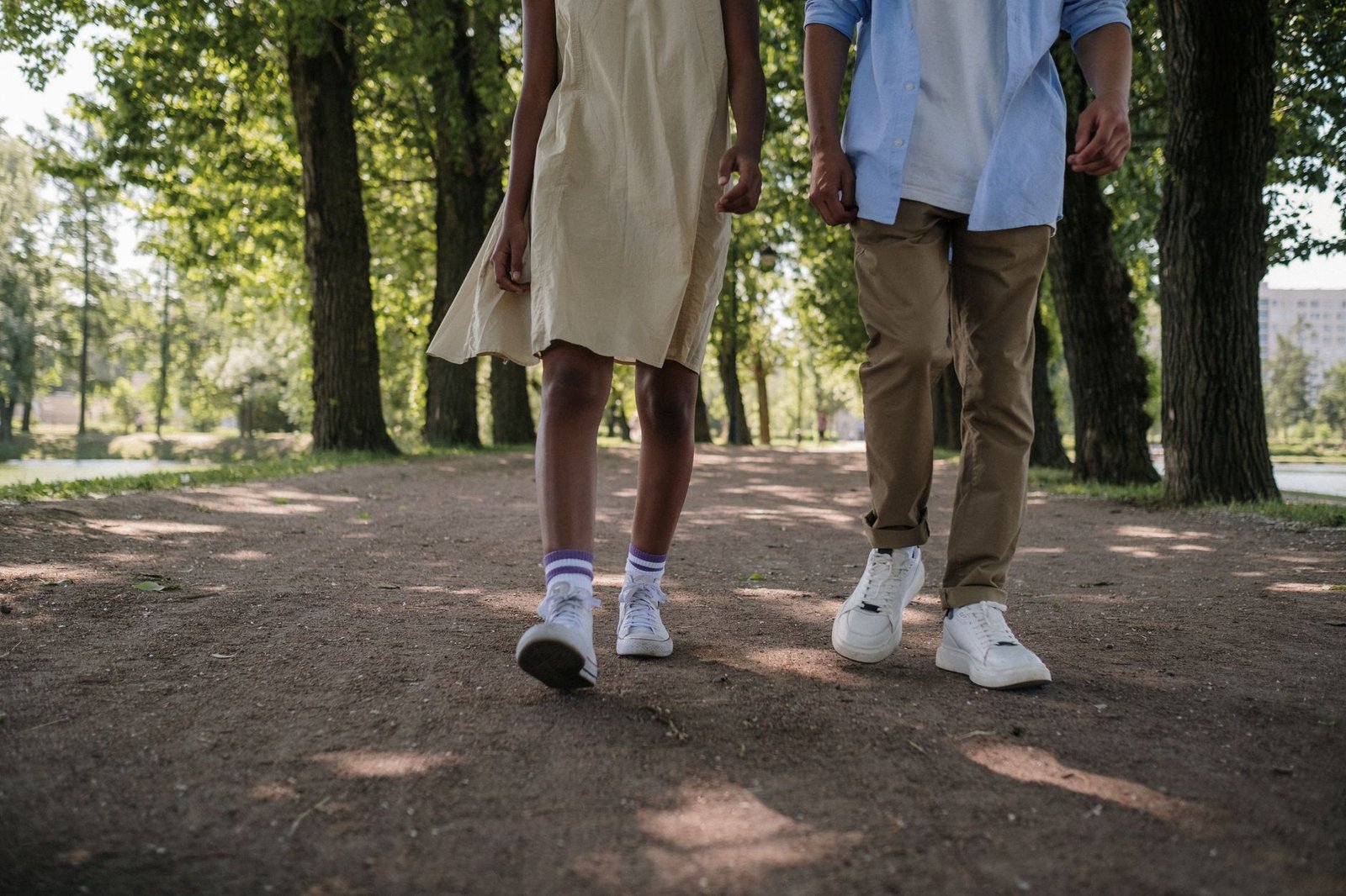 A teenage couple walks down a tree-lined path in a sunny park, evoking a sense of romance and tranquility.