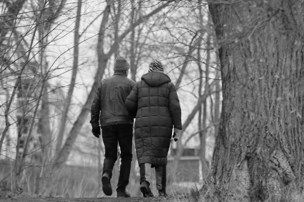 A couple walks arm-in-arm through a snowy winter forest, captured in black and white.