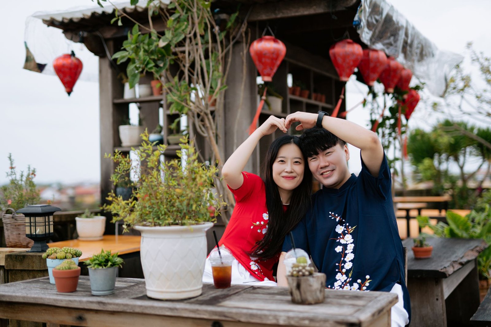Cheerful couple in floral shirts posing with a heart gesture in an outdoor café decorated with Chinese lanterns.