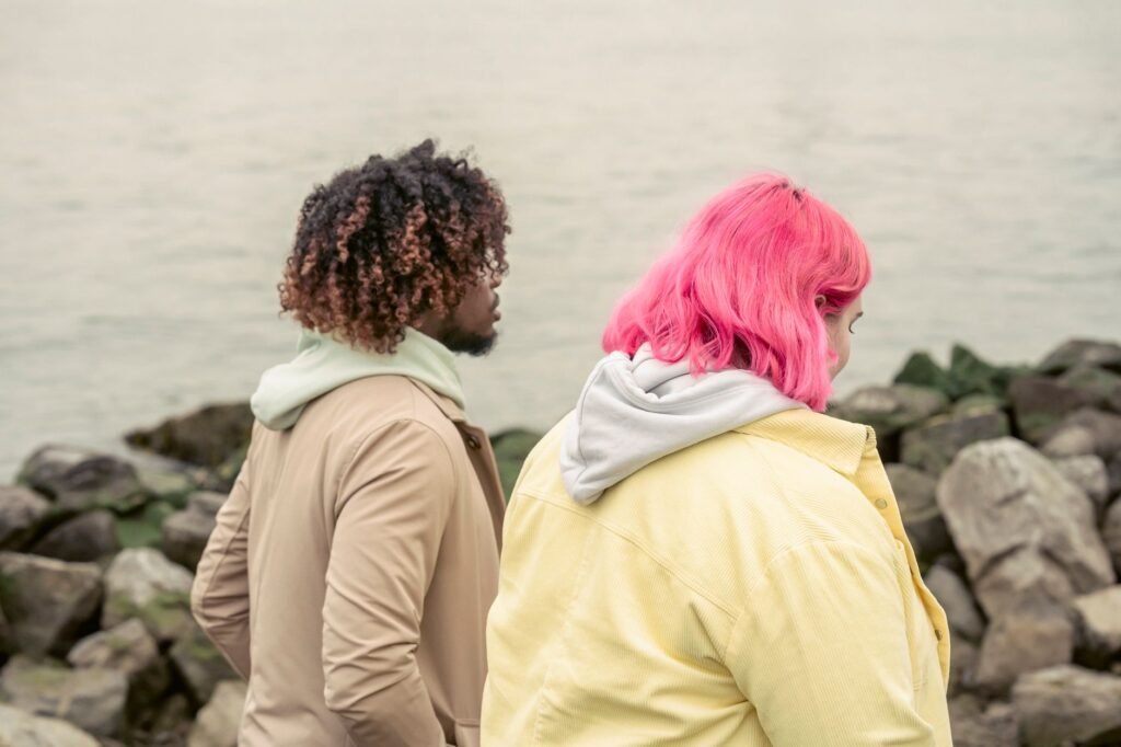 Side view of unrecognizable multiracial couple on outerwear strolling on waterfront with stones near calm water during date i