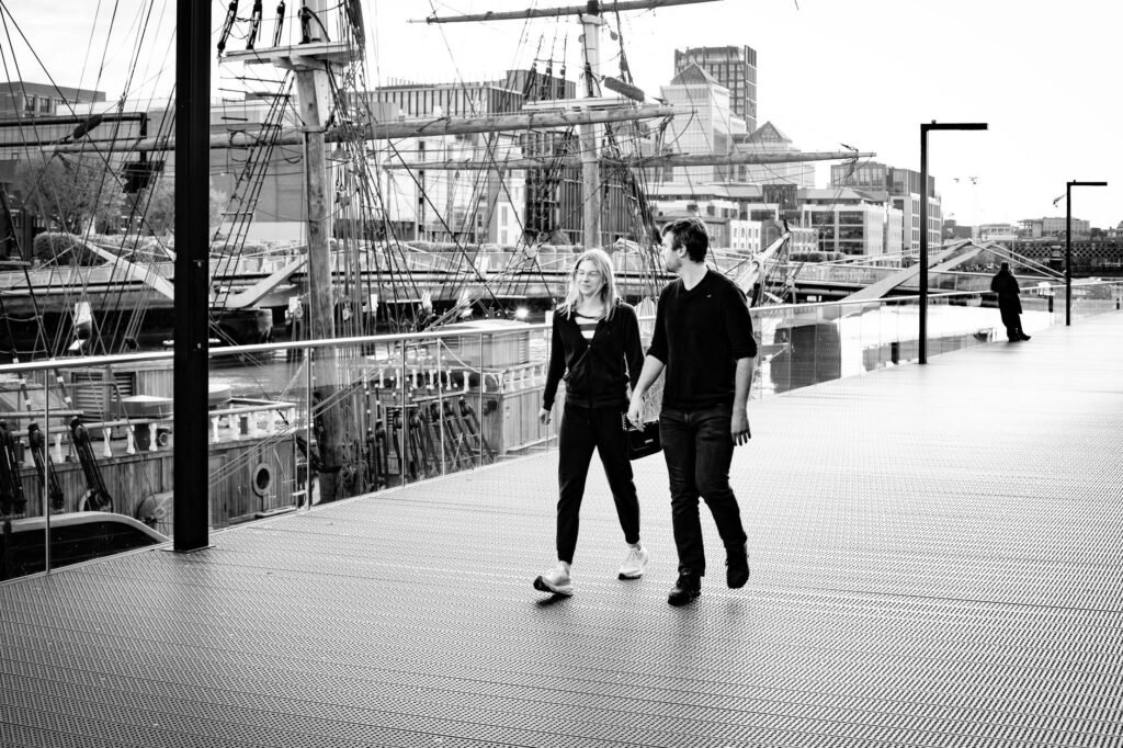 A couple walks along a Dublin dock, past a historic ship, in a lively urban setting.