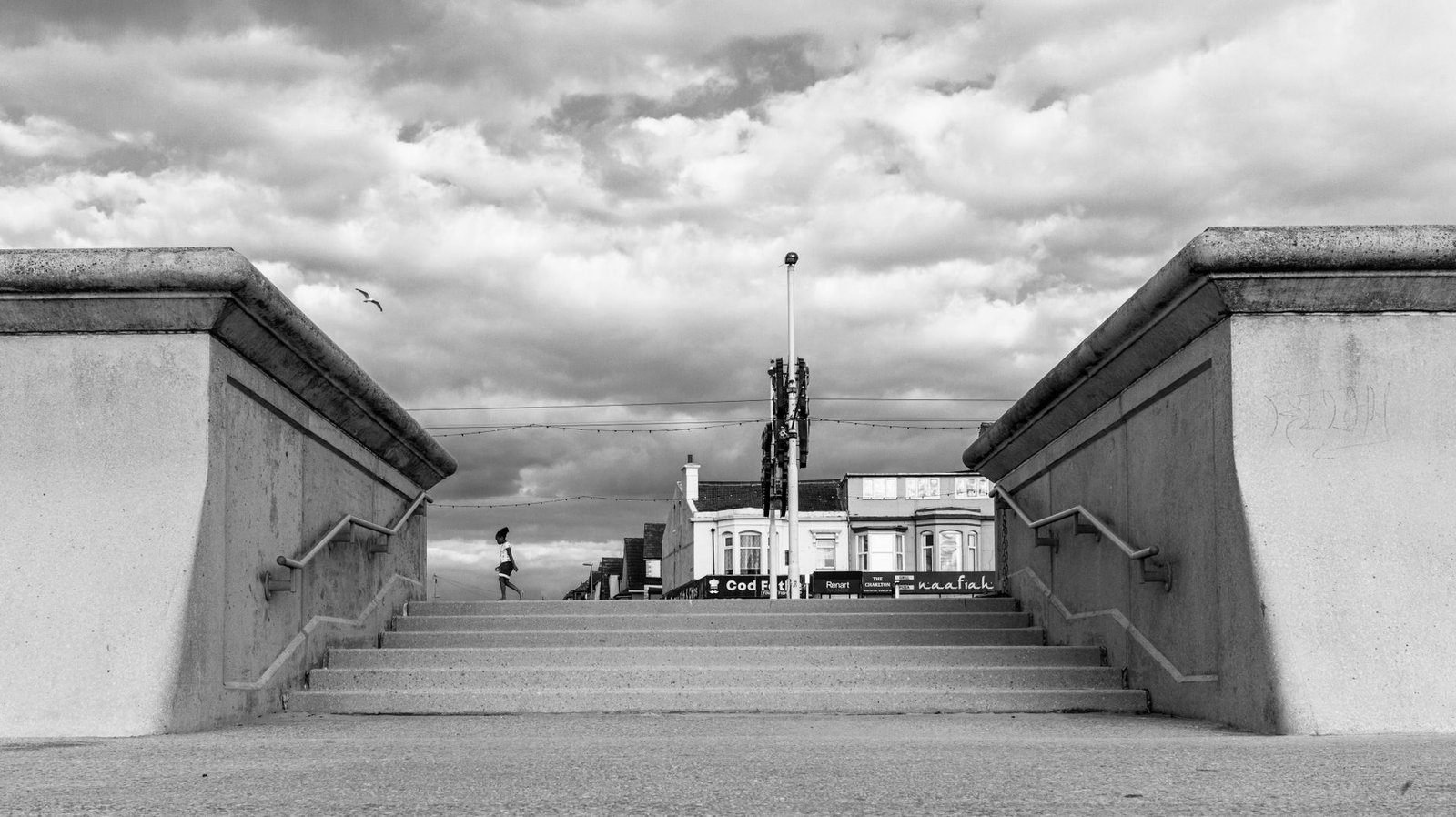 A monochrome image of a person walking by the stairs on Blackpool Promenade with a dramatic cloudy sky.