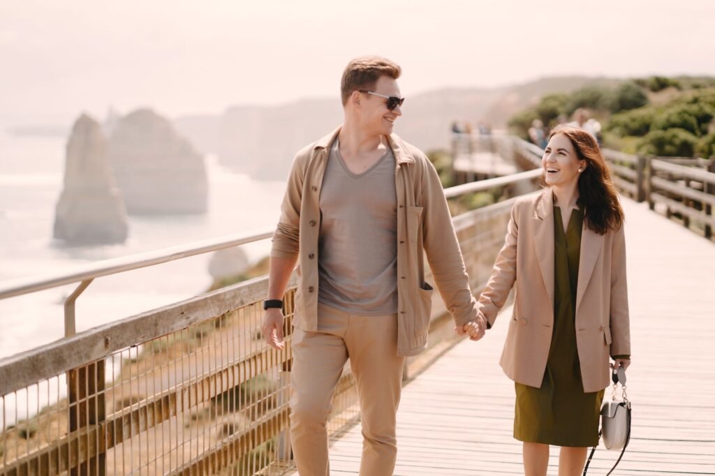 Positive smiling couple in stylish outfits holding hands and walking on promenade near waterfront against picturesque views o