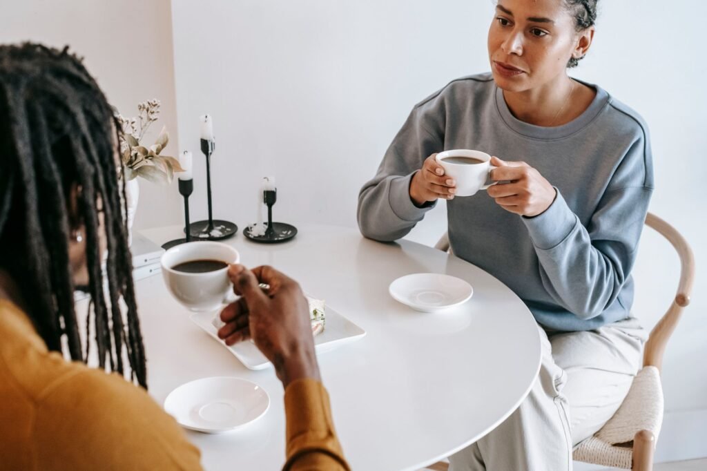 From above of crop pensive Hispanic female in casual clothes sitting at light table and drinking coffee with African American