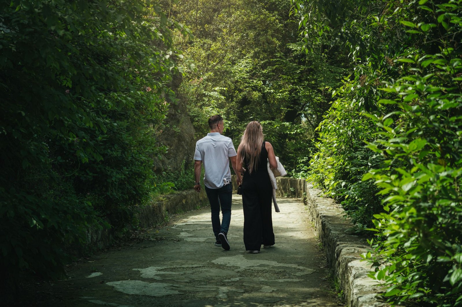 A couple walks hand in hand down a verdant forest path, surrounded by lush greenery.