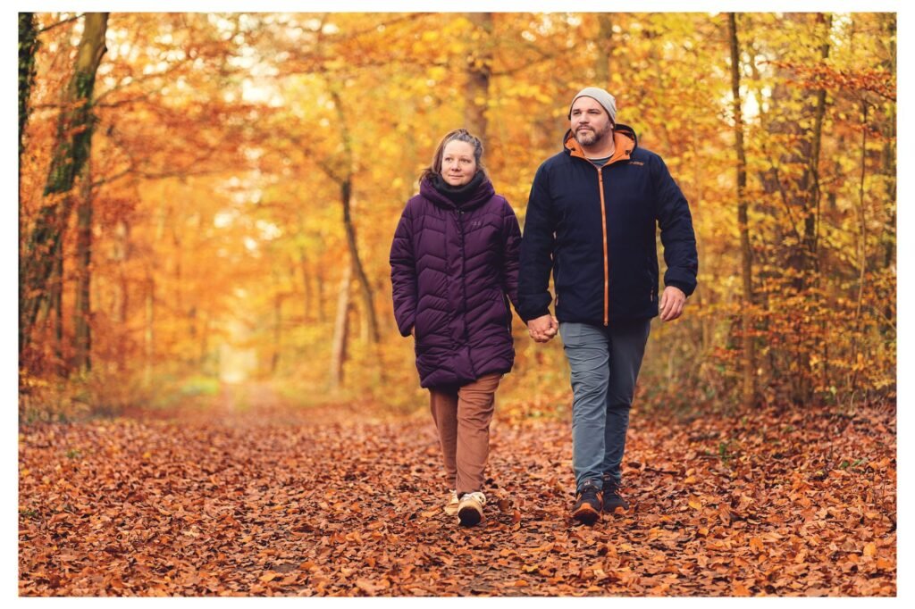 A couple walks hand in hand on a leaf-covered forest trail in autumn.