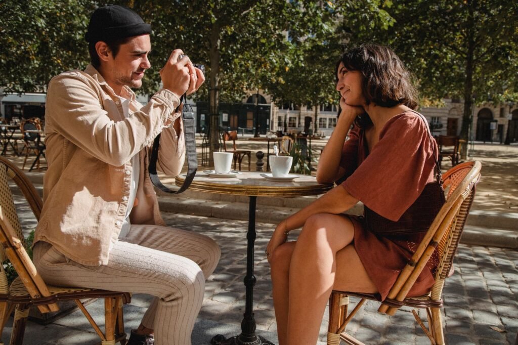 A couple enjoys a sunny day at an outdoor cafe, capturing the moment with a camera.