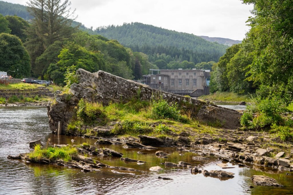 Tranquil river scene with rocky riverbanks, lush forestry, and historic dam in Scotland.