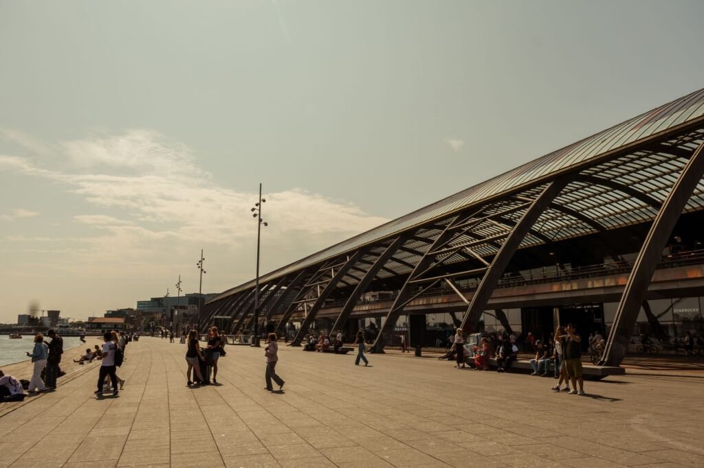 Wide walkway by Amsterdam Central Station, showcasing modern architecture and river view.