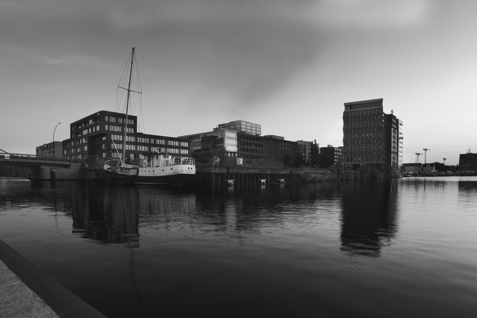 A serene black and white cityscape featuring a harbor with modern buildings and a sailboat reflection at twilight.