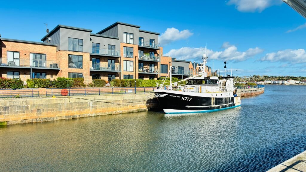 Contemporary apartments overlooking a peaceful quayside with a moored fishing boat under a bright blue sky.