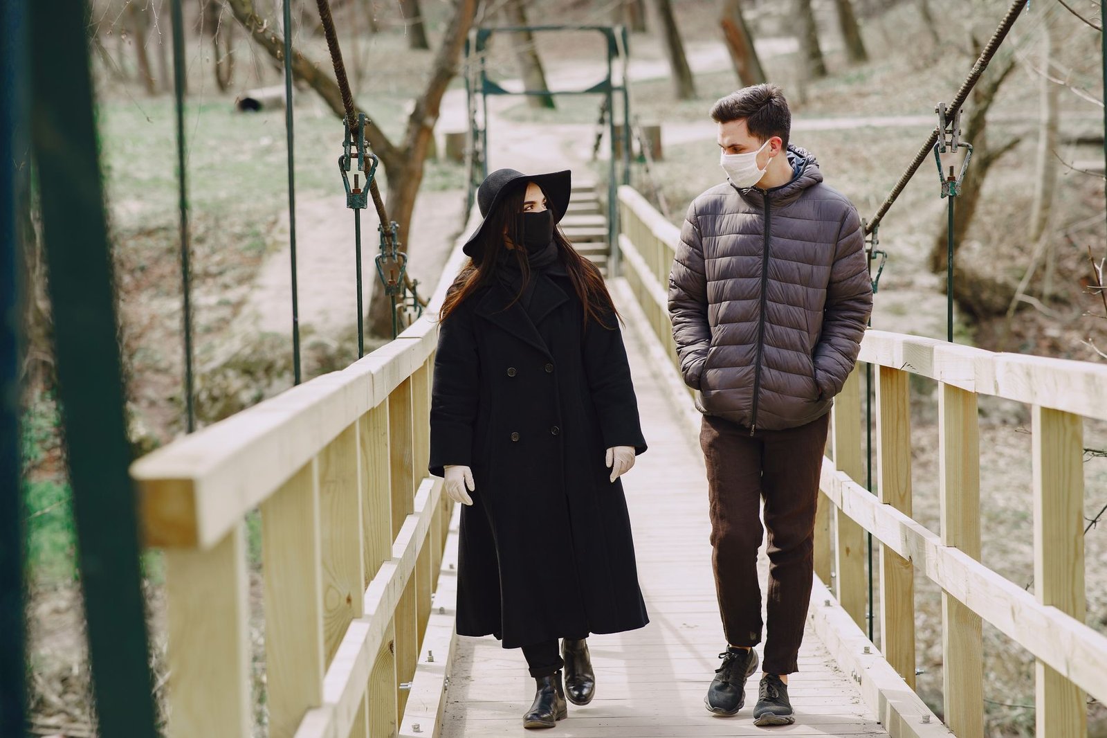 Full body young couple in outerwear and protective masks walking along wooden bridge in autumn park and talking to each other