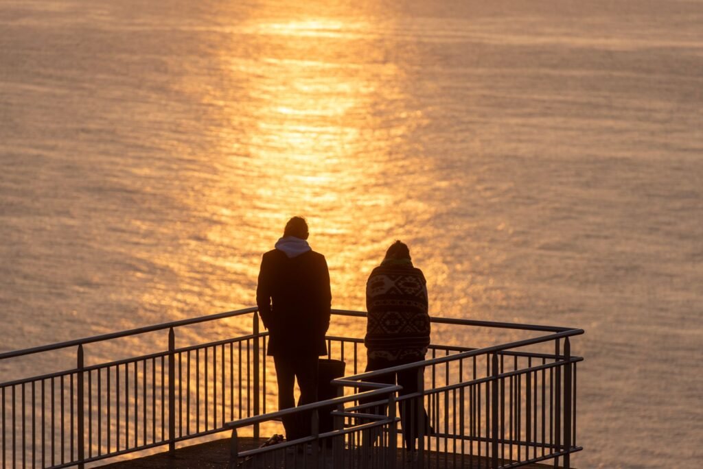 Two figures stand on a railing, silhouetted against a golden sea during sunset.