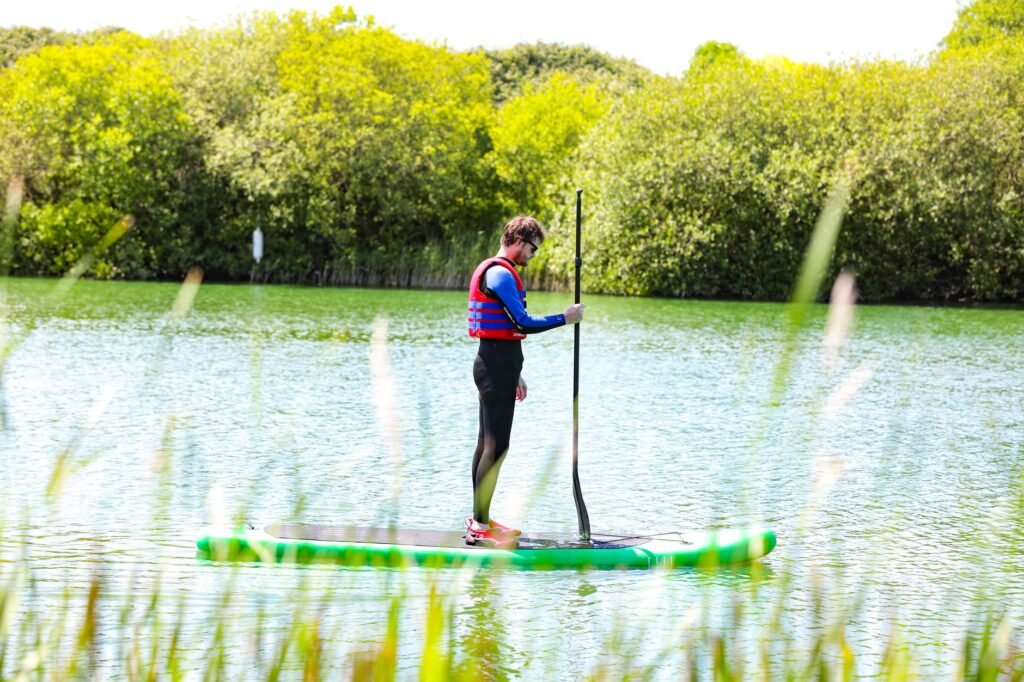 A man stands paddleboarding on a scenic river in England, wearing a life jacket under the bright day sky.