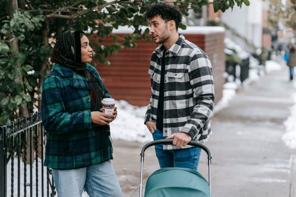 Ethnic woman wearing headscarf standing with cup of takeaway drink and talking to husband standing near stroller on snowy str