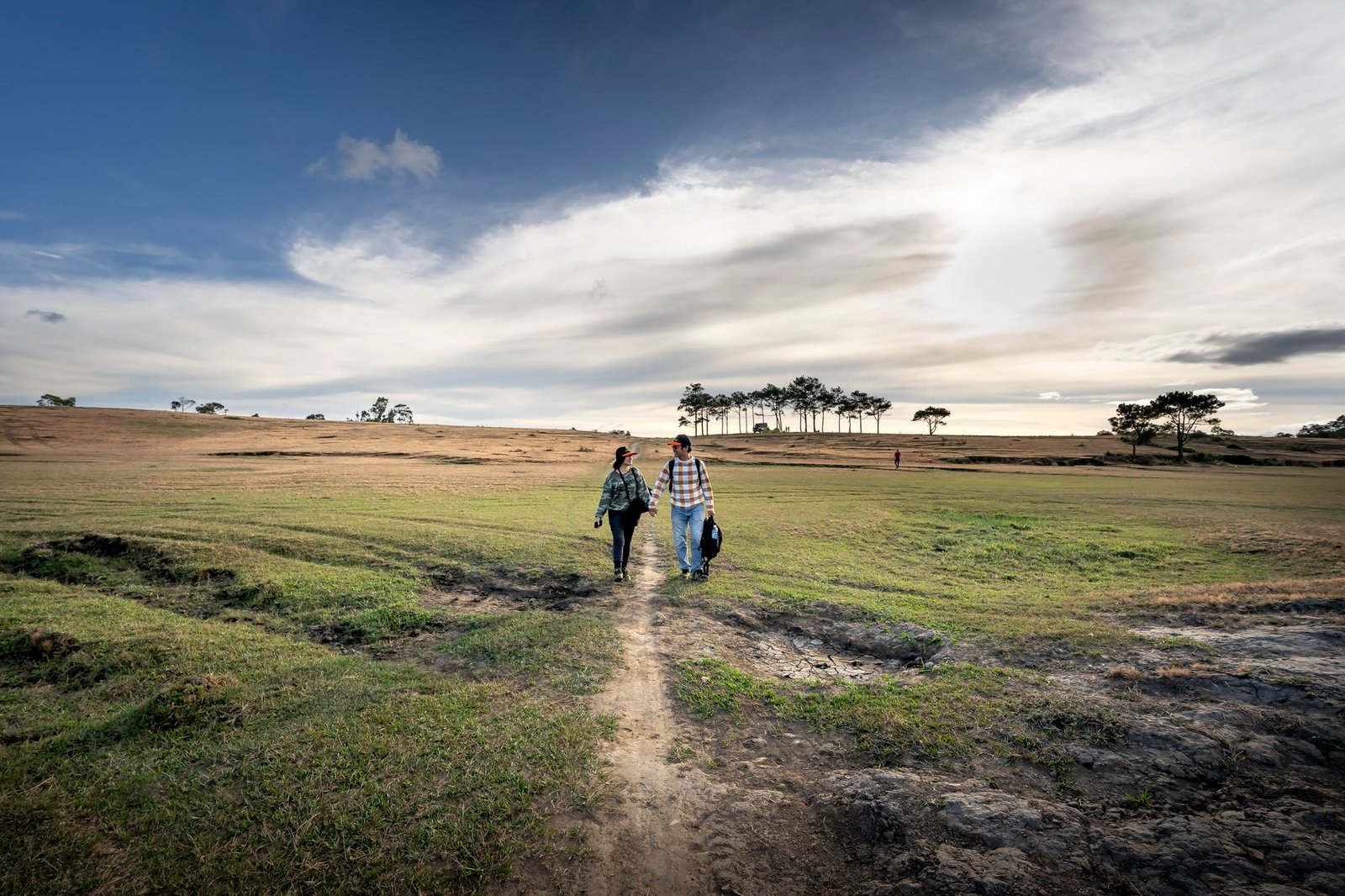 A couple holds hands while walking through a vast open field under a cloudy sky.