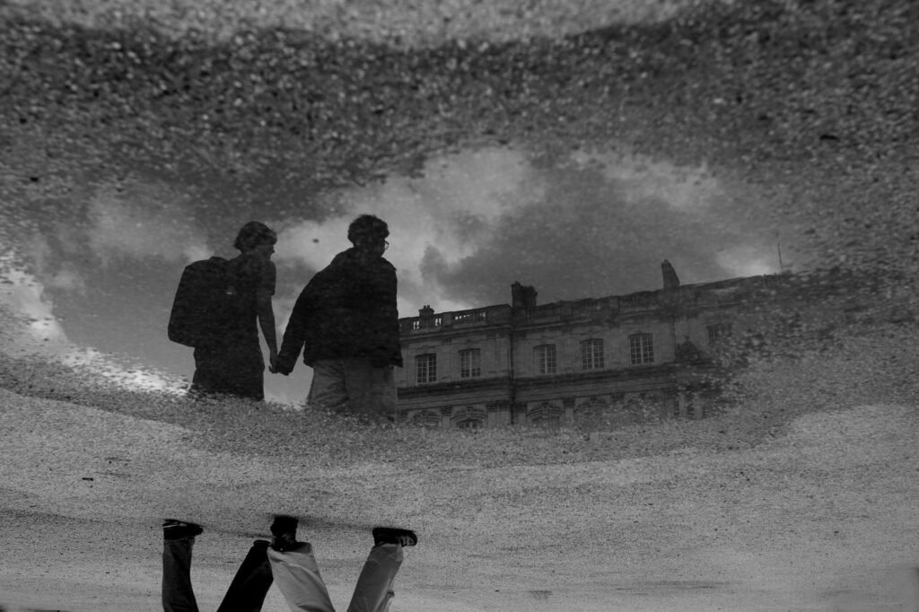 Reflection of a couple holding hands in a puddle by an historic building, captured in dramatic black and white.