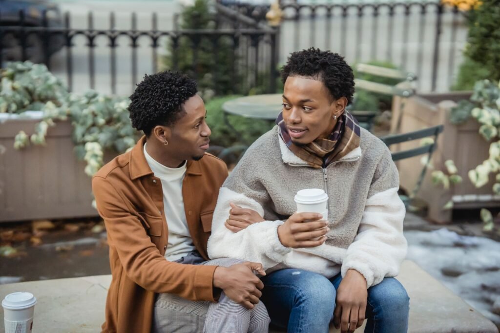 Positive African American homosexual couple with takeaway coffee looking at each other while sitting on bench during date on