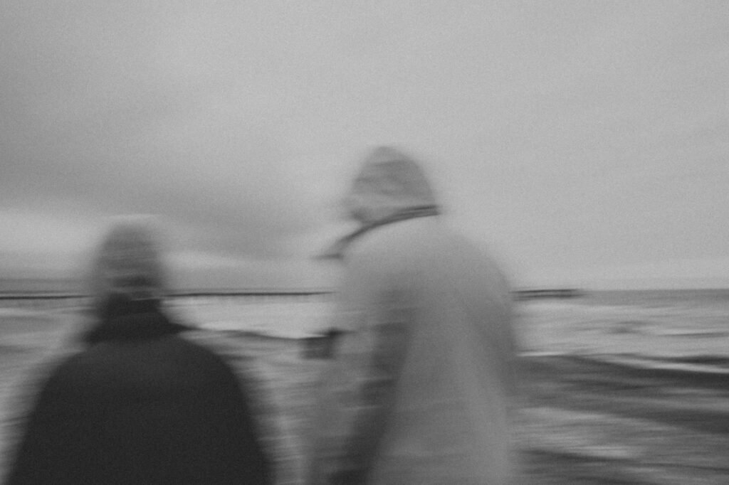 Artistic black and white photo of people walking on a cloudy beach with a blurred effect, evoking mystery.