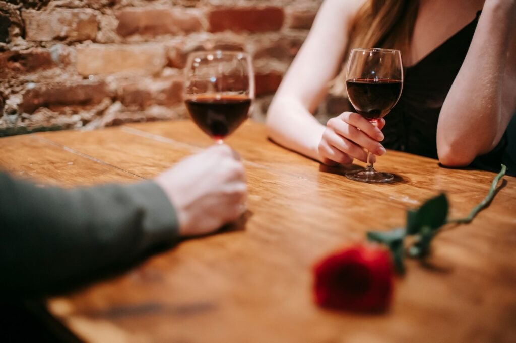 Crop anonymous couple in elegant outfits enjoying romantic date in bar at table with red rose and glasses with wine near bric