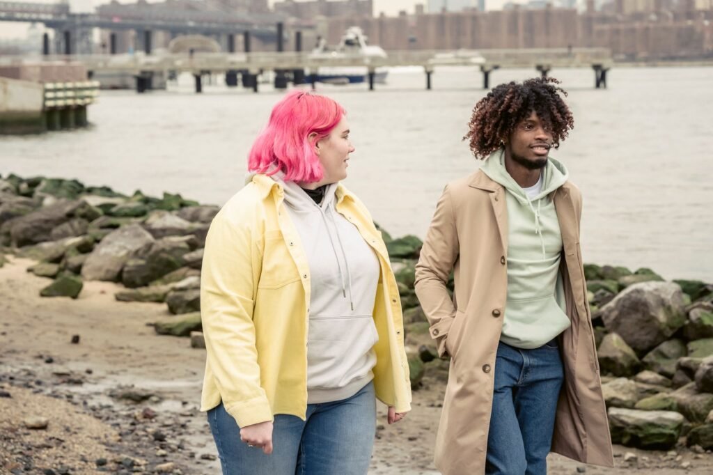 Content multiracial couple in outerwear strolling on embankment with stones near calm river against bridge and residential bu