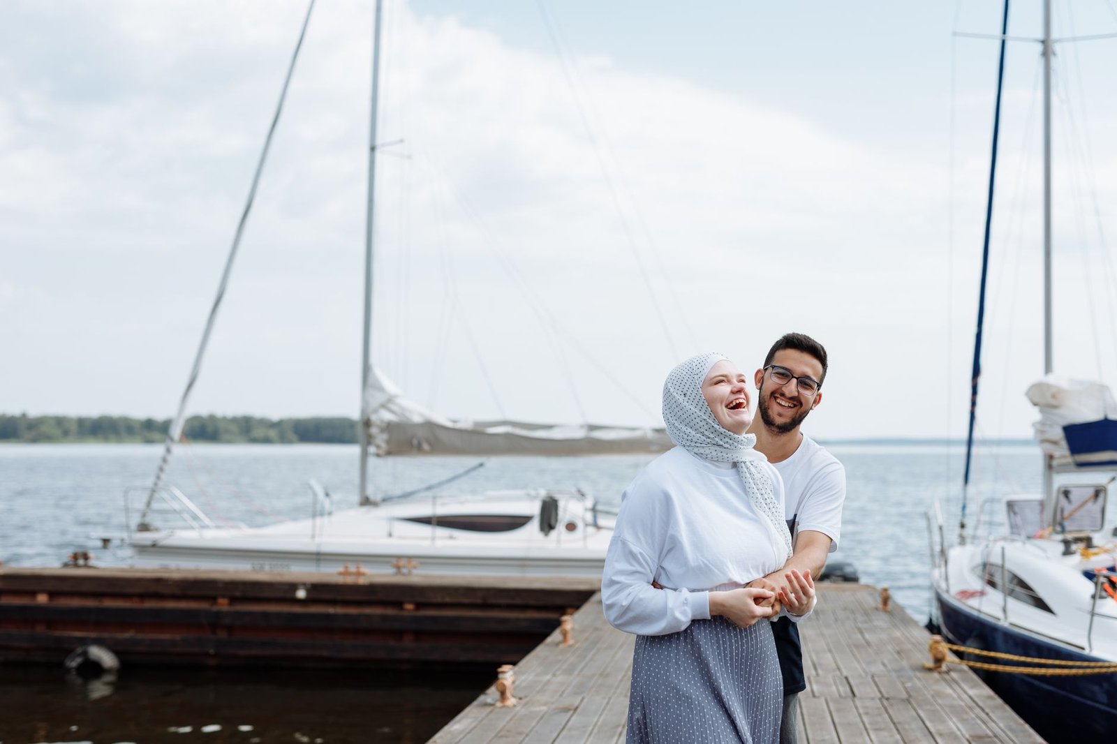 Joyful couple laughing together on a wooden dock by the marina, surrounded by boats and clear skies.